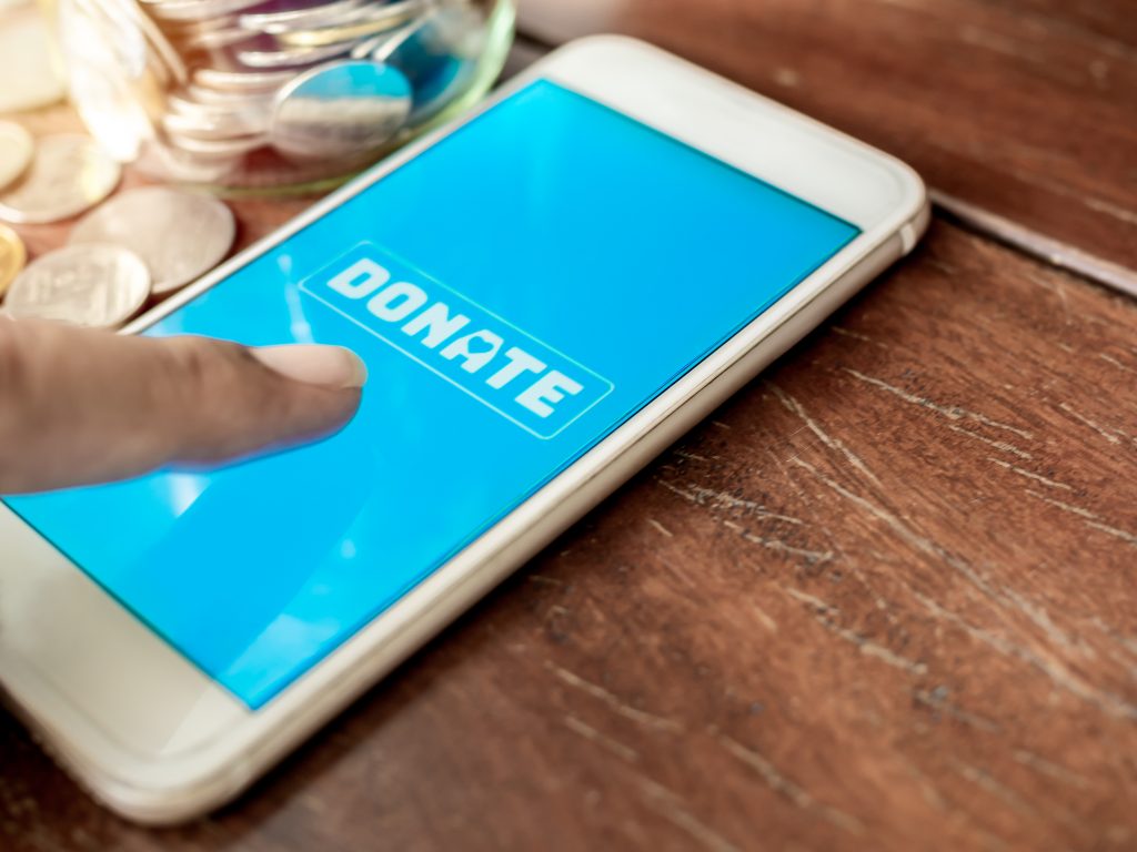 Finger pressing donate icon button on blue screen on white mobile phone with coins money on wooden background with copy space. Donation online concept.