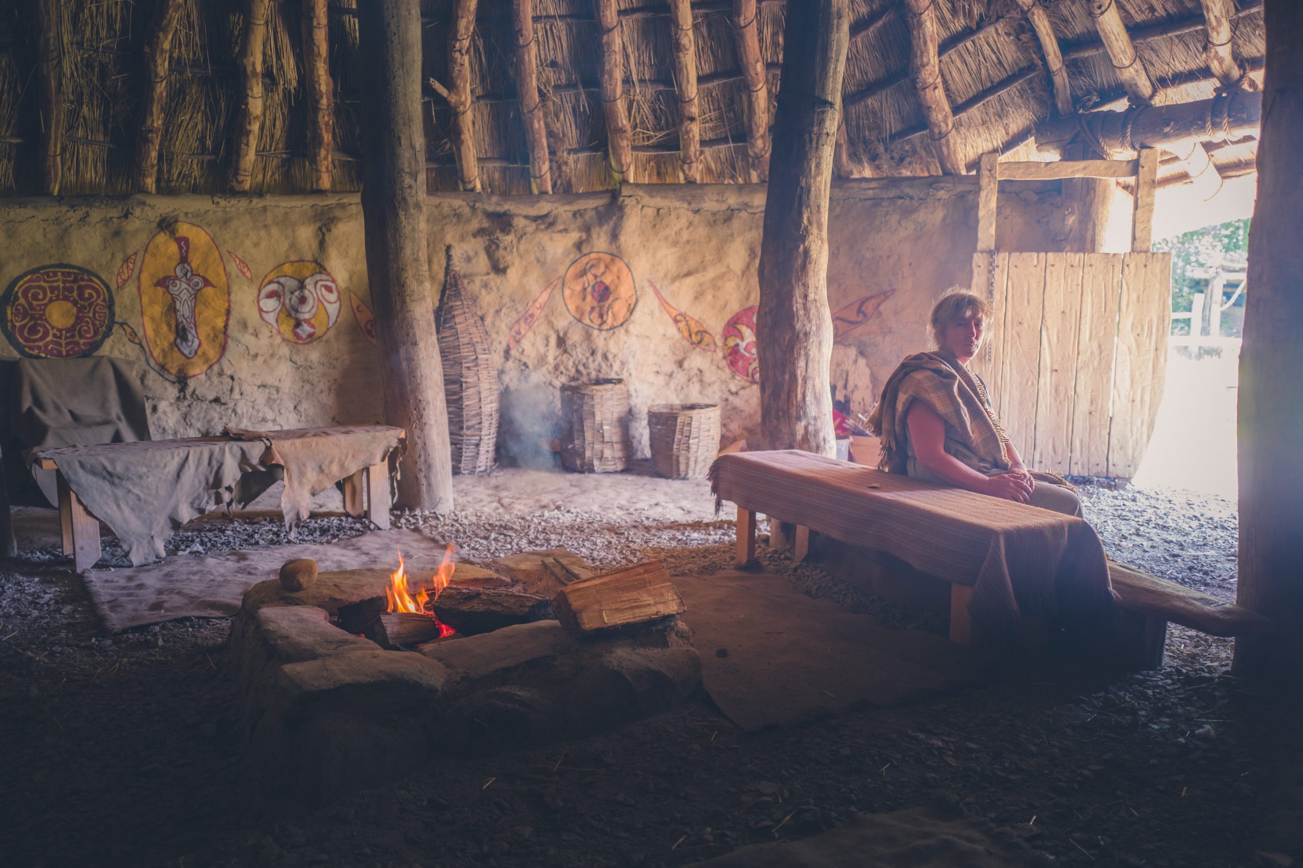 Woman dressed in Iron Age costume sitting inside roundhouse as campfire burns