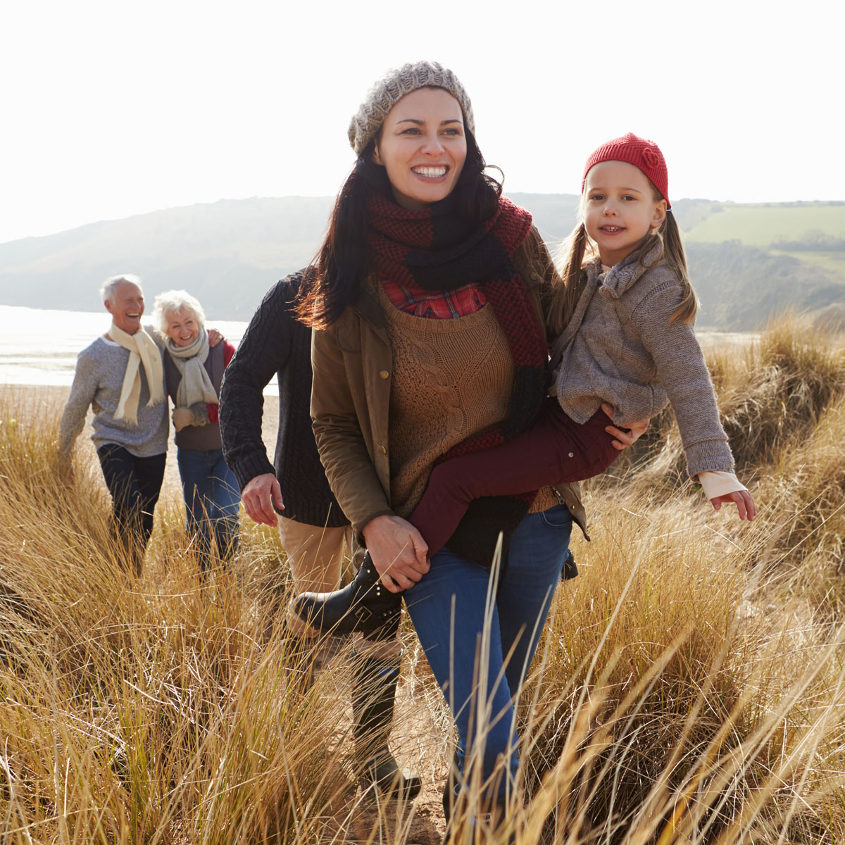 Multi generational family walking through long grassed sand dune beside beach