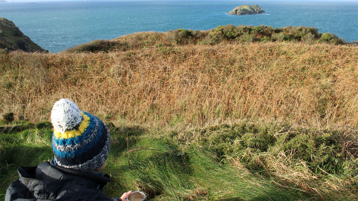 Person in bobble hat sitting on the grass looking out to see on a sunny day