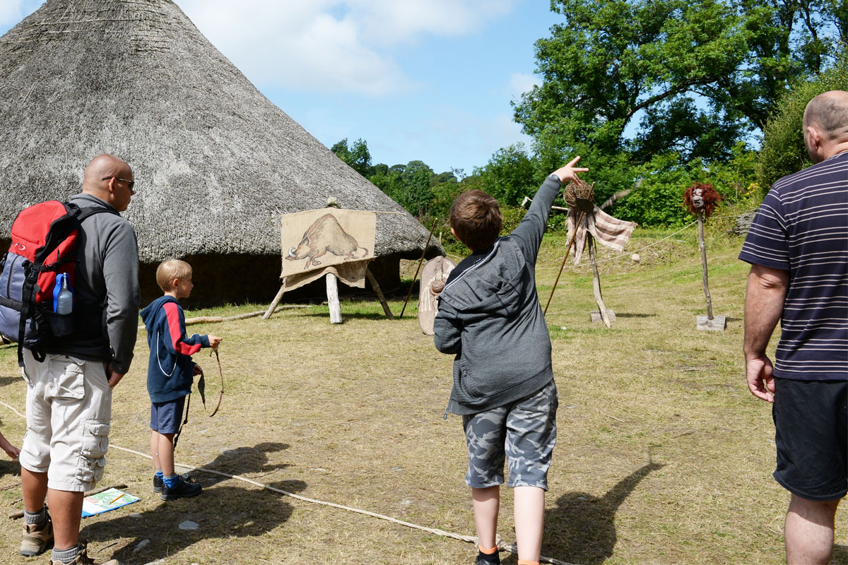 Group of people firing traditional slingshots outside a celtic roundhouse with a thatched roof