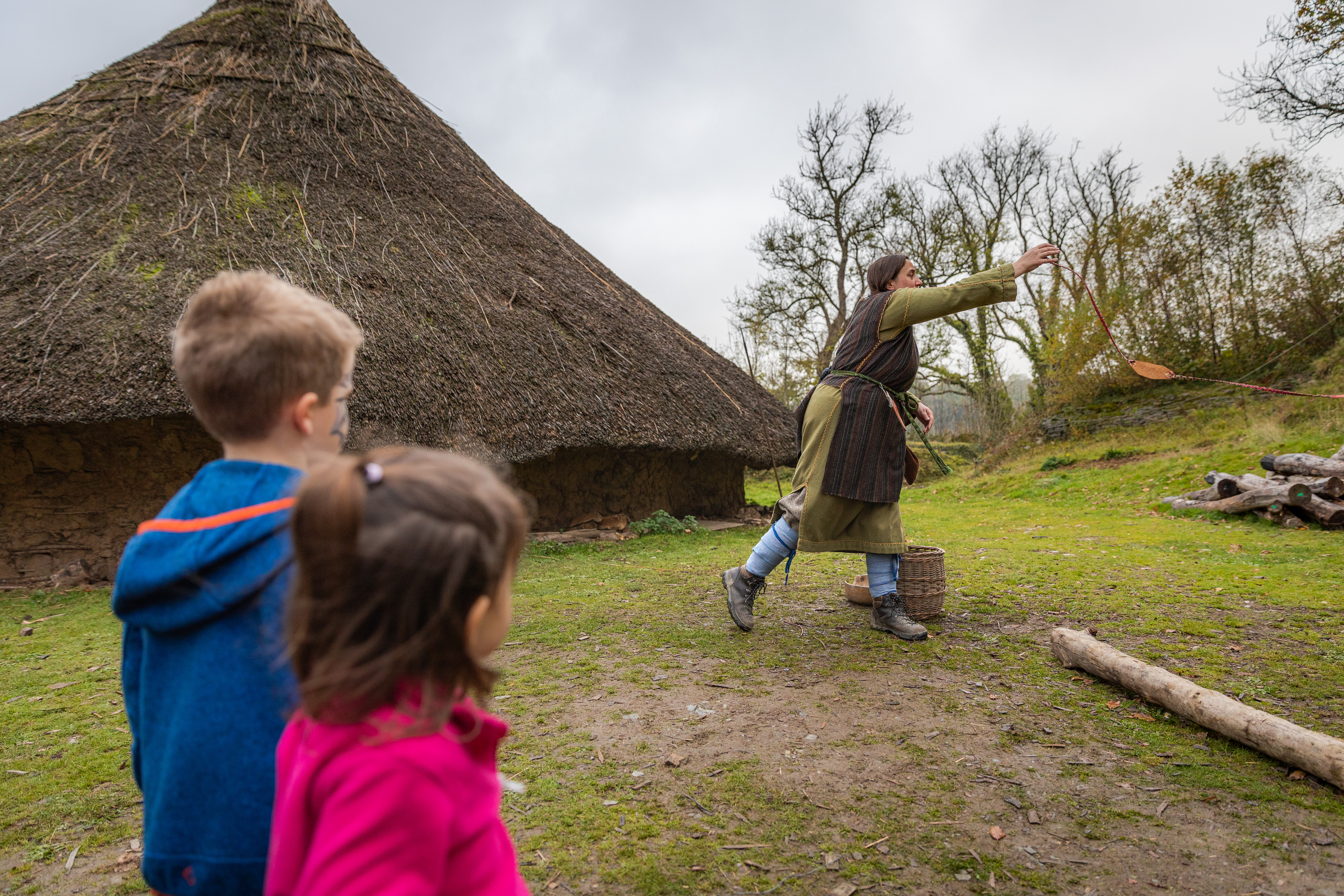 Two children watch a woman in celtic dress fire a traditional slingshot