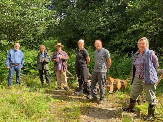 6 adults standing on grass in the wooded area with sawn logs behind them