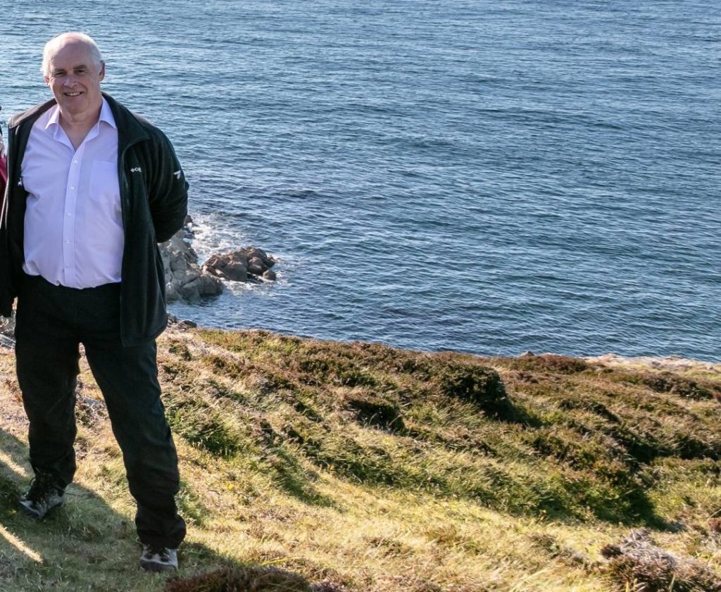 Man standing on a grassy cliff edge with a calm blue sea in the background