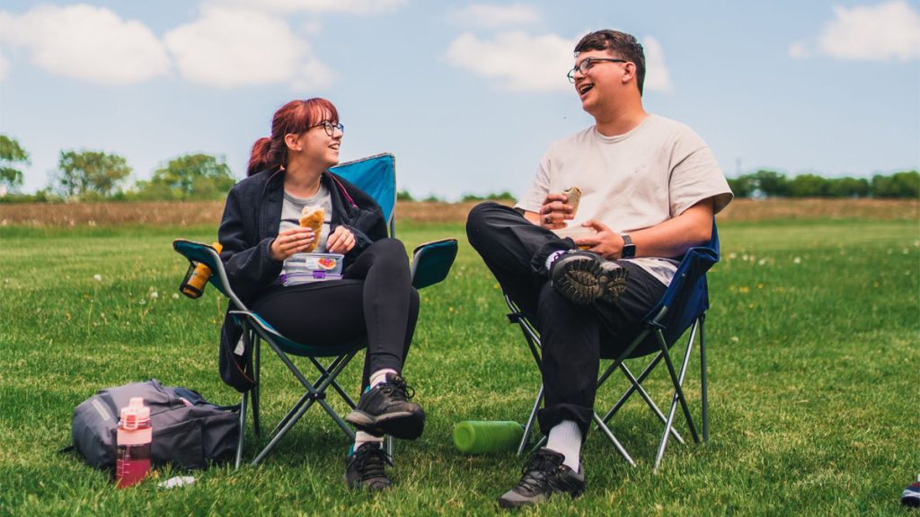 Woman and man sitting in camping chairs earing a picnic in a grassy campsite field