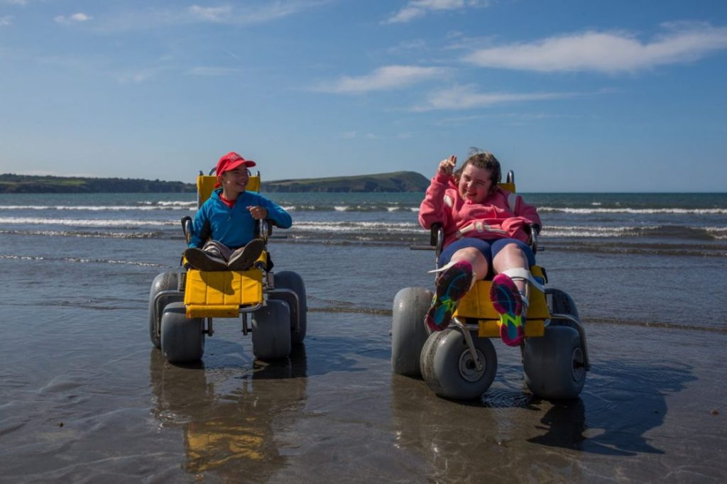 Two children on a beach using beach wheelchairs