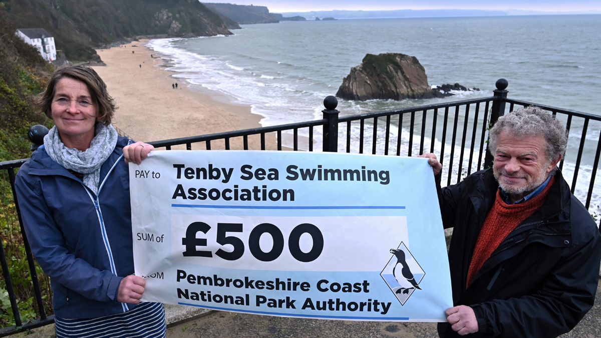 Two people holding a giant cheque aloft, standing above a sandy beach in winter. Cheque reads 'Tenby Sea Swimming Association £500 from Pembrokeshire Coast National Park Authority