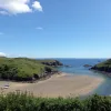 Sunny day photograph of an opening to a sandy harbour flanked by grassy cliffs either side. Location is Solva, Pembrokeshire.