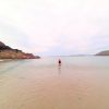 Christine Blumer Manorbier 2022 Single female in a one piece bathing suit standing in knee-high water at a sandy beach. Sandstone cliffs can be seen to the left and right. Location pictured is Manorbier, Pembrokeshire