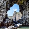 View of limestone cliffs through a hole in another limestone cliff across a rocky shore on a sunny day. Location pictured is Lydstep caverns, Pembrokeshire