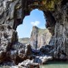 Guy Candler Lydstep Cavern 2014 View of limestone cliffs through a hole in another limestone cliff across a rocky shore on a sunny day. Location pictured is Lydstep caverns, Pembrokeshire