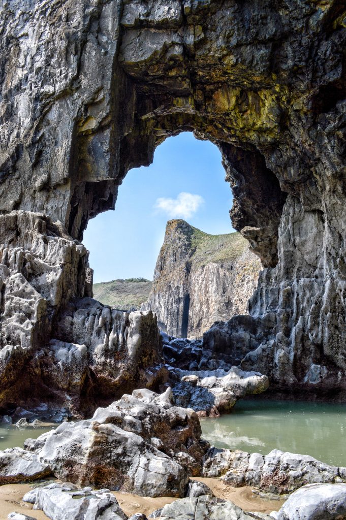View of limestone cliffs through a hole in another limestone cliff across a rocky shore on a sunny day. Location pictured is Lydstep caverns, Pembrokeshire