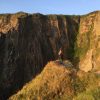 Hannah Gover Between Pwll Deri and Aberbach 2021 Single person standing on the edge of a grassy headline looking out across a void to rocky cliffs opposite. Location is between Pwll Deri and Aberbach on the Pembrokeshire Coast Path