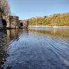 Keith Solva Harbour 2022 Two lime kilns located on the left hand side of a harbour with boats moored in the distance and a wooded cliff behind with houses perched atop the cliff. Photograph is taken across the shallow water at low tide. Location pictured is Solva, Pembrokeshire