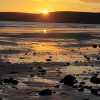 View across a sandy beach at sunset littered with stones scattered across the shore. The sun is setting behind the cliffs in the distance creating an orange sky, which is reflecting in the waters left behind on the shore by the receding tide. Location pictured is Newgale, Pembrokeshire