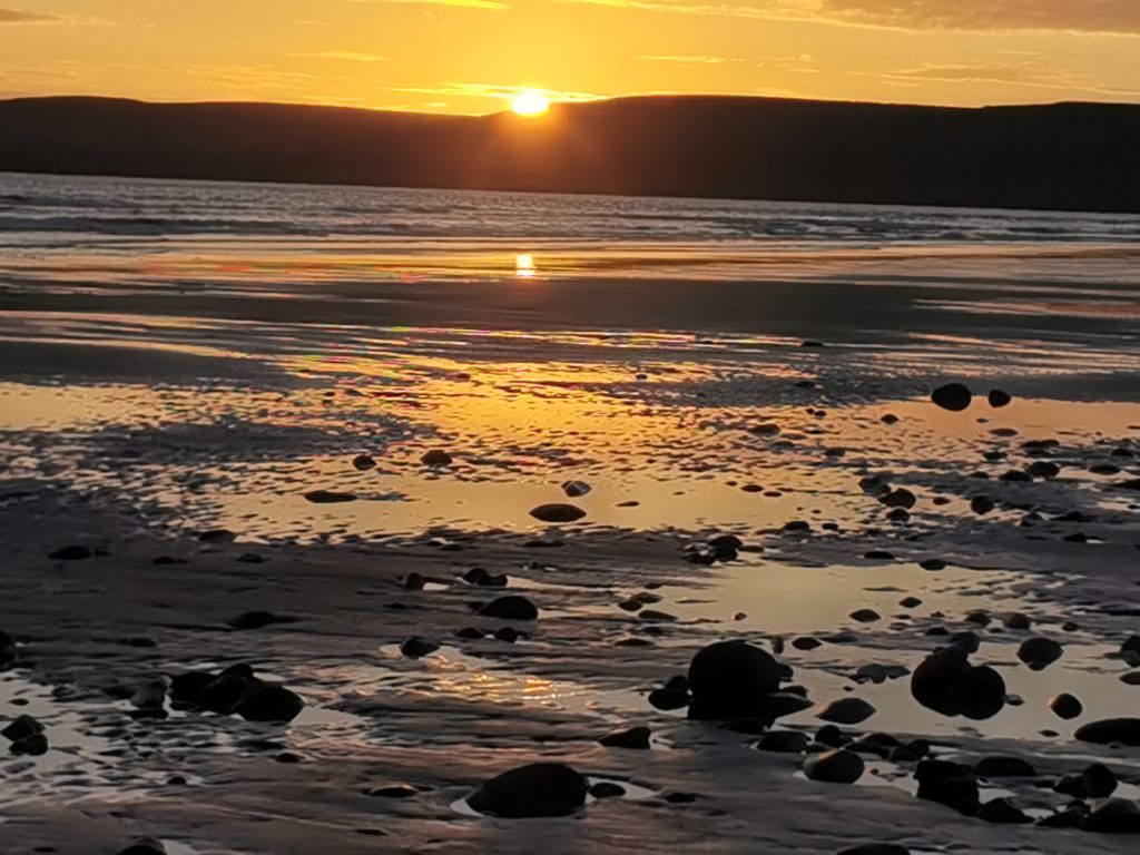View across a sandy beach at sunset littered with stones scattered across the shore. The sun is setting behind the cliffs in the distance creating an orange sky, which is reflecting in the waters left behind on the shore by the receding tide. Location pictured is Newgale, Pembrokeshire