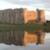 Mark Raikes Carew 2012 Partially ruined stone castle reflecting in the millpond it overlooks on an overcast day with the orange of a setting sun reflecting on one side of the castle walls. Location featured is Carew Castle, Pembrokeshire