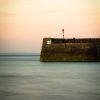 Rhys Walker Tenby 2022 Image of a stone harbour wall with light blue sea and orangey sky. Location pictured is Tenby, Pembrokeshire
