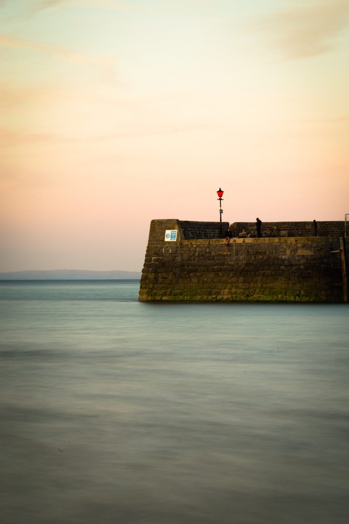 Image of a stone harbour wall with light blue sea and orangey sky. Location pictured is Tenby, Pembrokeshire