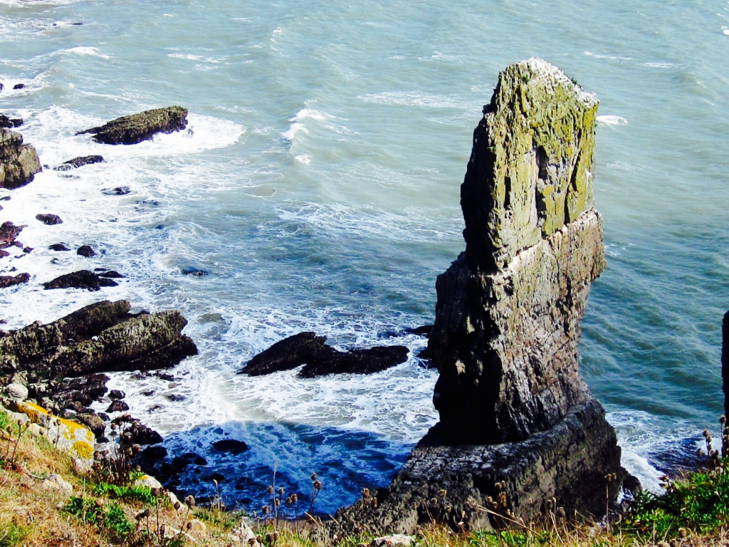 Limestone stack jutting out of the sea next to a grassy cliff edge with rough seas lapping the shore