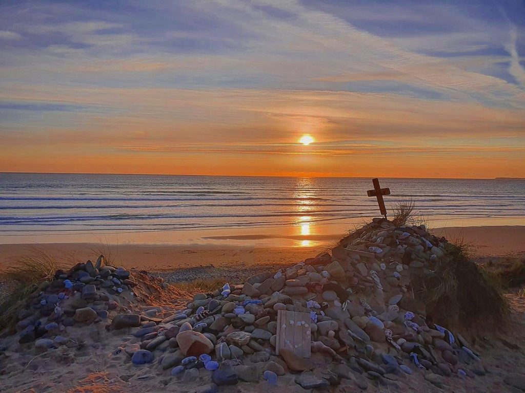 The sun setting above a sandy beach creating an orange glow across the sky and sea. In the foreground a pile of pebbles has been added to small sand dune mound with a small crucifix as its top. This is to mark the apparent site of Dobby&#039;s Grave, a fictional character from the Harry Potter books.