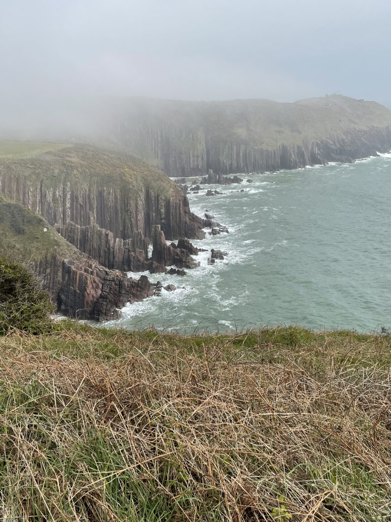 Photograph of limestone cliffs topped with grass on a misty day with bluegreen seas crashing against the bottom of the cliffs.