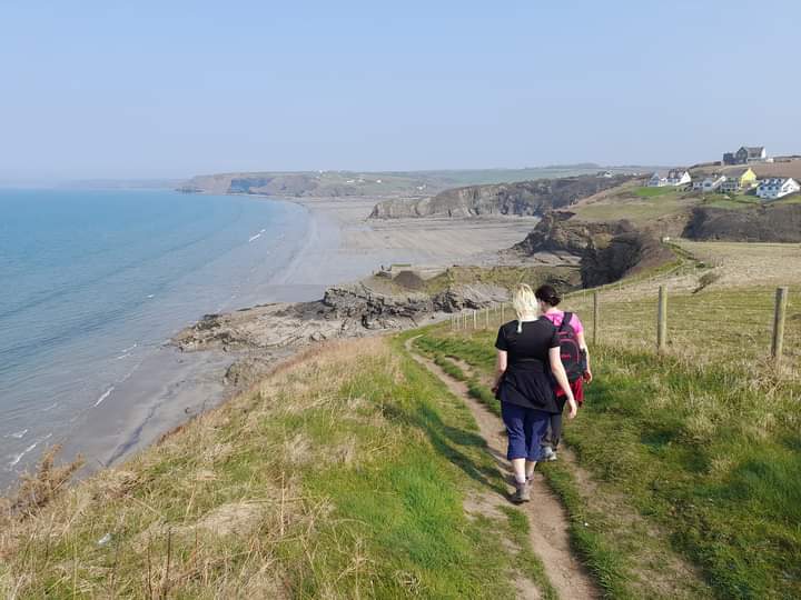 Two people walking along a grassy coastal footpath with rugged cliffs and sandy beaches in the distance and four houses on the cliffs to the right, with the sea to the left.