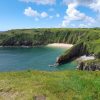 Sandy beach flanked by grassy rugged cliffs taken on a sunny day with only a few fluffy white clouds in the blue sky. Location pictured is Skrinkle Haven, Pembrokeshire