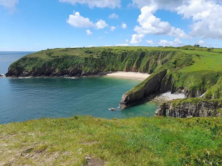Sandy beach flanked by grassy rugged cliffs taken on a sunny day with only a few fluffy white clouds in the blue sky. Location pictured is Skrinkle Haven, Pembrokeshire