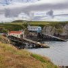 Simon Carpenter St Justinian Lifeboat Station 2020 Two lifeboat stations located in a rocky cove amongst grass-covered cliffs. One is an older style boat house with cream sides and a curved red roof, the other is more modern with pastel blue sides and a grey roof. Location pictured is St Justinian, Pembrokeshire