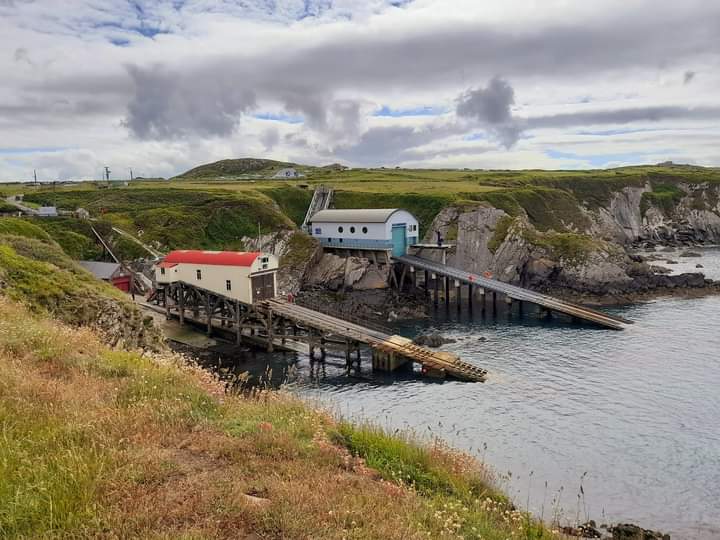 Two lifeboat stations located in a rocky cove amongst grass-covered cliffs. One is an older style boat house with cream sides and a curved red roof, the other is more modern with pastel blue sides and a grey roof. Location pictured is St Justinian, Pembrokeshire
