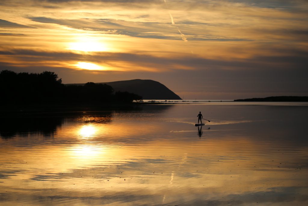 Single stand up paddleboarder floating on a calm sea as the sun sets creating an orange glow in the sky, which is reflected on the sea. The paddleboarder is in silhouette, as are the cliffs either side of the mouth of the estuary leading to the sea. Location pictured is the Nevern Estuary, Newport, Pembrokeshire