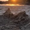 Mounds of sand on a beach with a sunset glowing orange in the background out of focus, reflecting on the sea as it laps the shore. Location pictured is Angle, Pembrokeshire