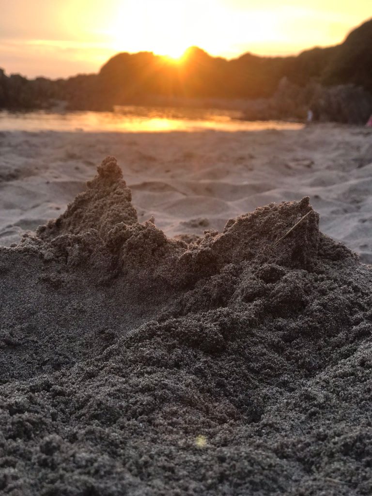Mounds of sand on a beach with a sunset glowing orange in the background out of focus, reflecting on the sea as it laps the shore. Location pictured is Angle, Pembrokeshire