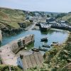 Photograph of a small fishing village with manmade harbour walls enclosing an area where several small boats are moored. Many cars are parked along the edge of the harbour wall and in the village behind it. Location pictured is Porthgain, Pembrokeshire