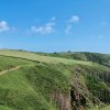 Timy Lyn Skrinkle 2022 Person walking along a coastal footpath which cuts a line along a grassy clifftop with limestone cliffs below. The sky is blue with very few small clouds floating in it. Location pictured is Skrinkle, Pembrokeshire
