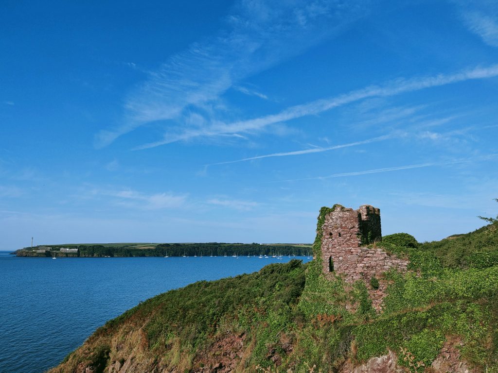 Ruined stone building on the edge of a cliff covered with grass and ivy with a view across to wooded coastline in the background with a number of boats moored next to the cliff. It&#039;s a sunny day with blue skies and blue seas. Location pictured is St Anne&#039;s Head, Pembrokeshire