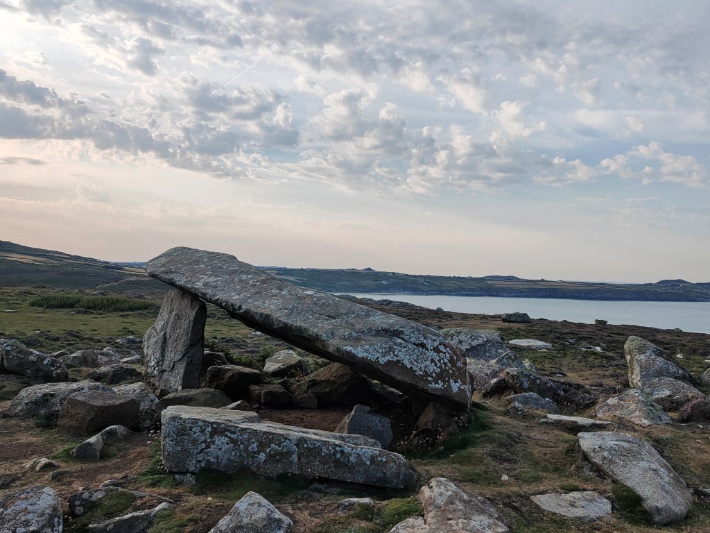 Remains of a stone burial chamber with one capstone being supported by two standing stones, one taller than another. There are a number of stones scattered around it and a view across the sea to a coastline in the background. Location pictured is Coetan Arthur on St David&#039;s Head, Pembrokeshire