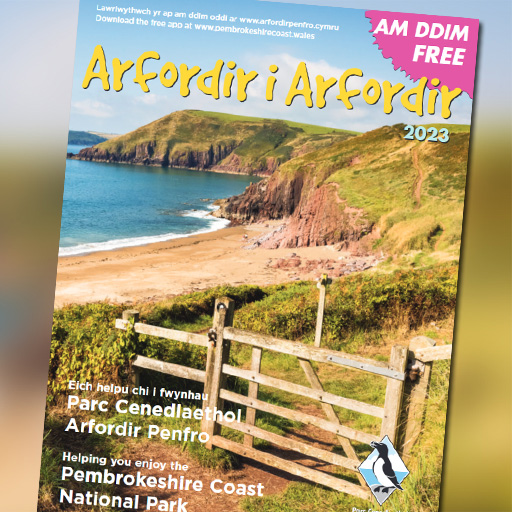 Cover of a magazine titled 'Arfordir i Arfordir' which features a coastal scene including a wooden gate across a footpath leading to a sandy beach with red sandstone cliffs in the distance.
