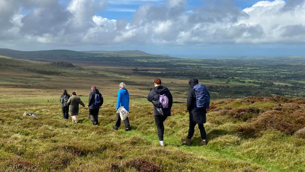 Six people walking on a grassy footpath towards a range of sweeping hills.