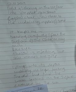 Image of a poem written on lined paper. It reads: Still seas. Light glistening on the surface like dappled gemstones. Crystal clear view down to the undulating ripples of sand. It tempts me in, treading carefully I feel the surface of the water on my skin Like silk, the cold is refreshing, but still makes me gasp  I plunge into its depths instantly no longer feeling the cold but find the warm sections on the surface caressed by the Sun.