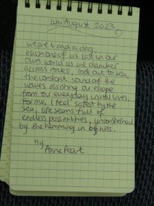 Poem written in a notebook. It reads: 4th August 2023. We are 6 and a dog Each one of us lost in our own world as we clamber across rocks, look out to sea,  The constant sound of the waves sound of the waves assisting our escape from our everday lives. For me, I feel safest by the sea, life seems full of endless possibilities, unconstrained by the hemming in of hills. By Anne Peart