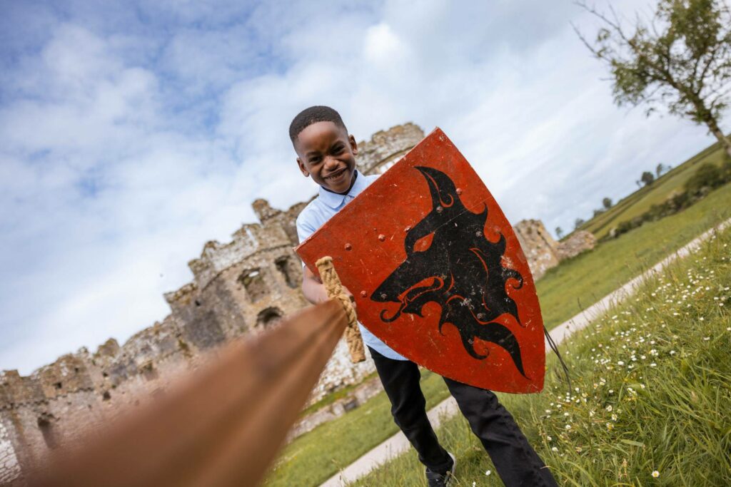 A young boy wielding a sword and shield, standing on front of Carew Castle.
