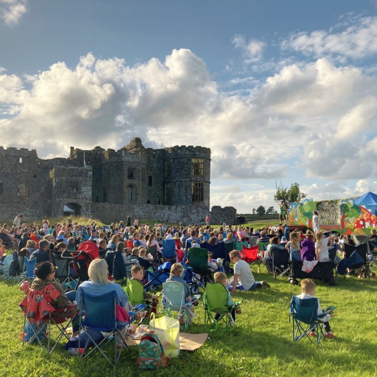 People seated outside on a fine summer's evening, watching an outdoor theatre performance, with Carew Castle in the background